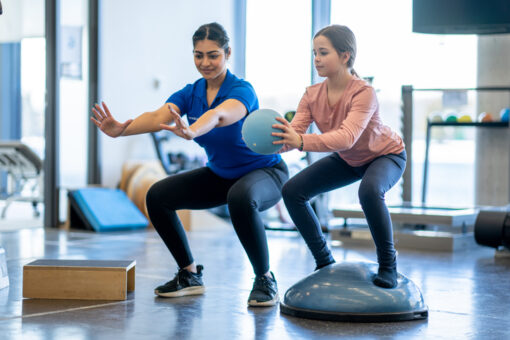 A young girl works with her physiotherapist in a gym during an appointment. She is dressed comfortably and is balancing on a half ball as she follows the therapist instructions and does a squat with a ball in her hands.